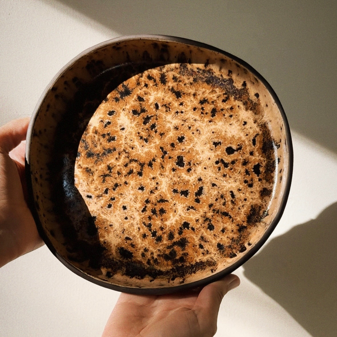 A handmade ceramic bowl with a speckled brown and black glaze, held from below by two hands against a warm beige wall with soft diagonal shadows. Made with the Fin Pottery Tools Irregular Round Blate Forms.