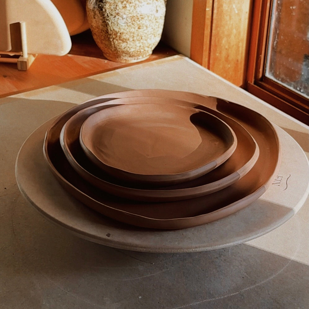 A stack of three nested ceramic plates made with warm brown clay, arranged on a bat. Made using the Fin Pottery Tools Irregular Round Plate Bundle.
